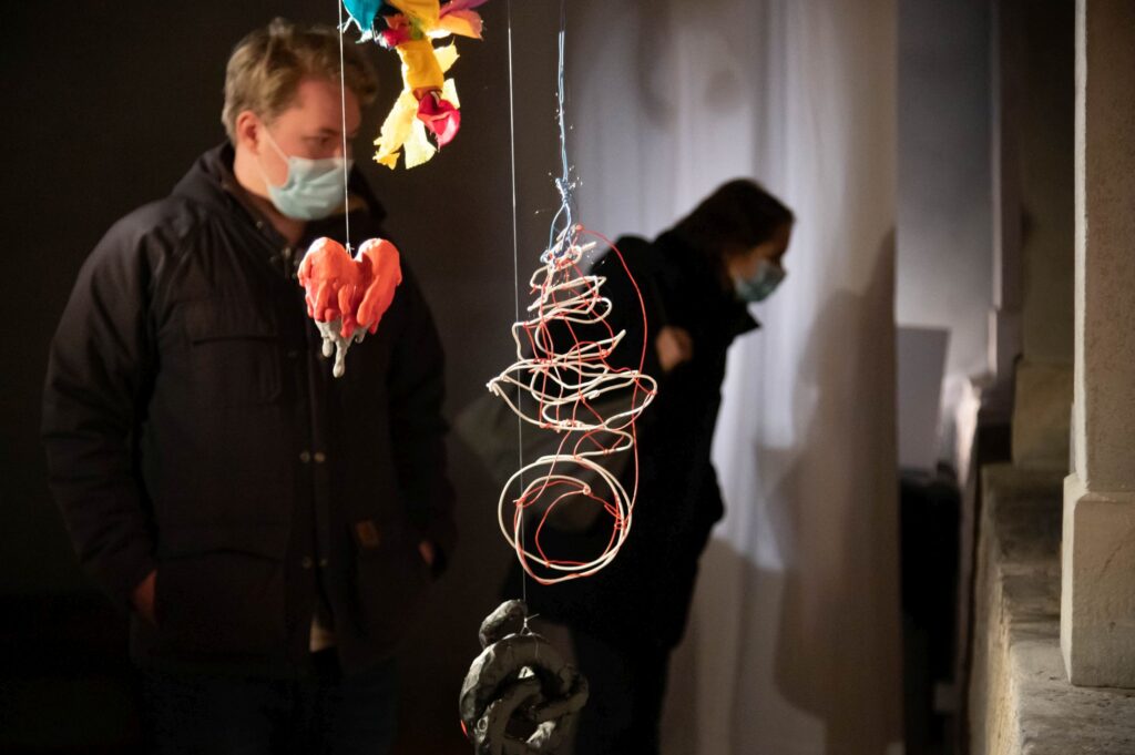 Skulptur von einem Herz, Geschwür und Faden. Im Hintergrund schauen zwei Menschen mit Maske die Ausstellung an.