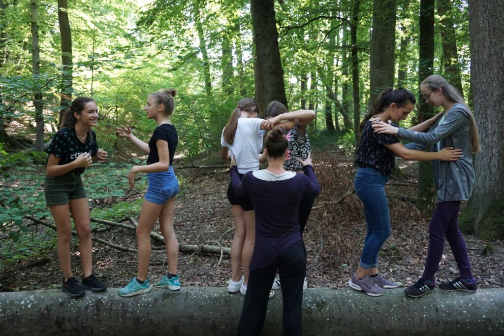 Junge Frauen tanzen jeweils zu zweit auf einem Baumstamm im Wald.