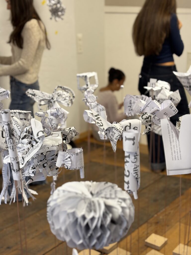 Weisse Papierskulpturen mit Text im Vordergrund, im Hintergrund Schülerinnen beim Aufbau der Skulpturen.