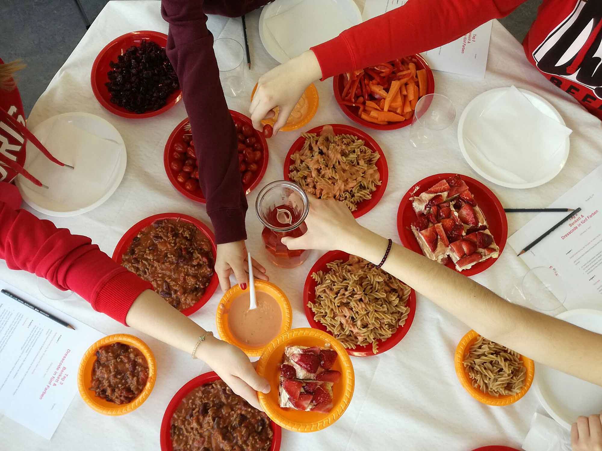 Speisen in roten und orangen Tellern aufgetischt, danach greifende Hände