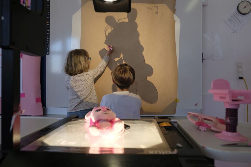 Zwei Kinder zeichnen den Schatten eines Plüschhasen nach, der von einem Hellraumprojektor an die Wand geworfen wird.