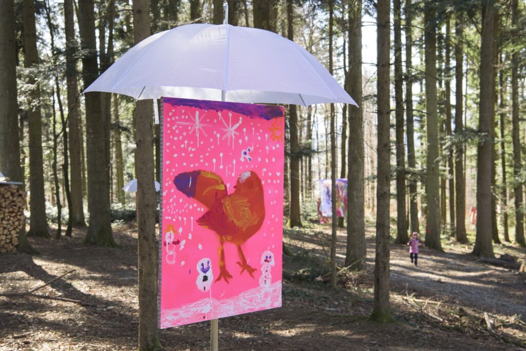 Kunstwerk von einem Kind steht unter einem weissen Regenschirm mitten im Wald.