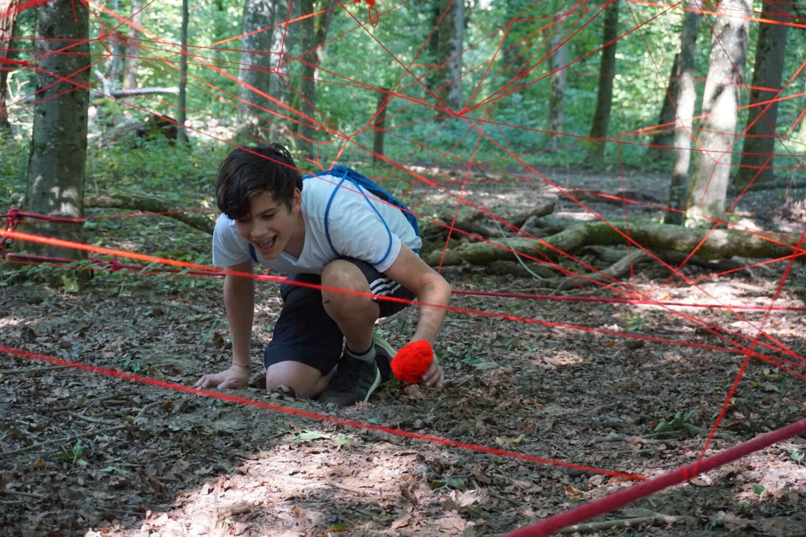 Jugendlicher kniet im Wald am Boden und spinnt mit rotem Faden ein Netzt.