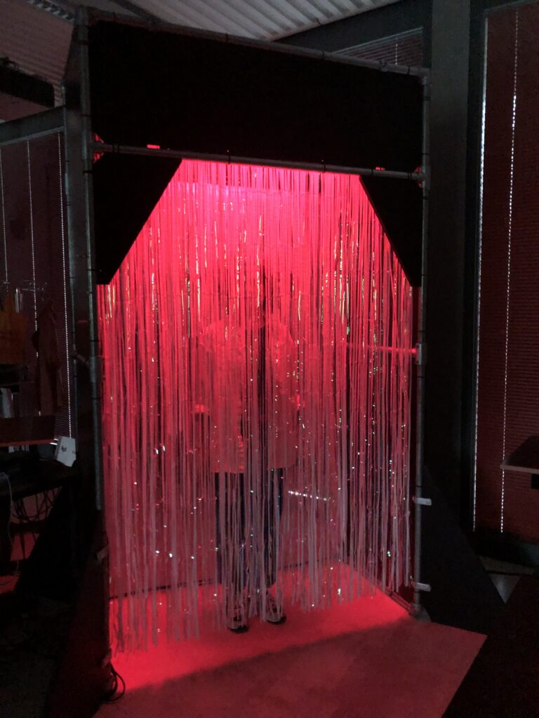 Rauminstallation mit dicht herabhängenden transparenten Fäden, die in rotes Licht getaucht sind.