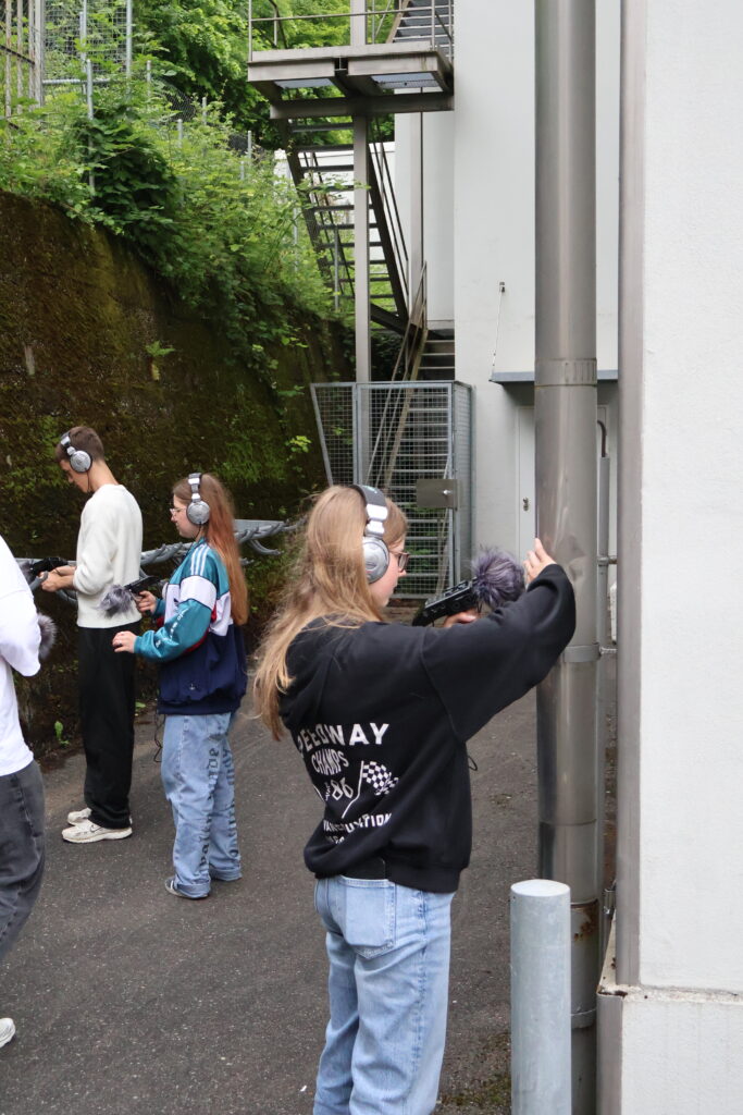 Junge Personen halten im aussenraum Aufnahmegerät und Mikrofon an verschiedene Objekte. Eine davon hält das Gerät an das Abflussrohr von einem Dach.