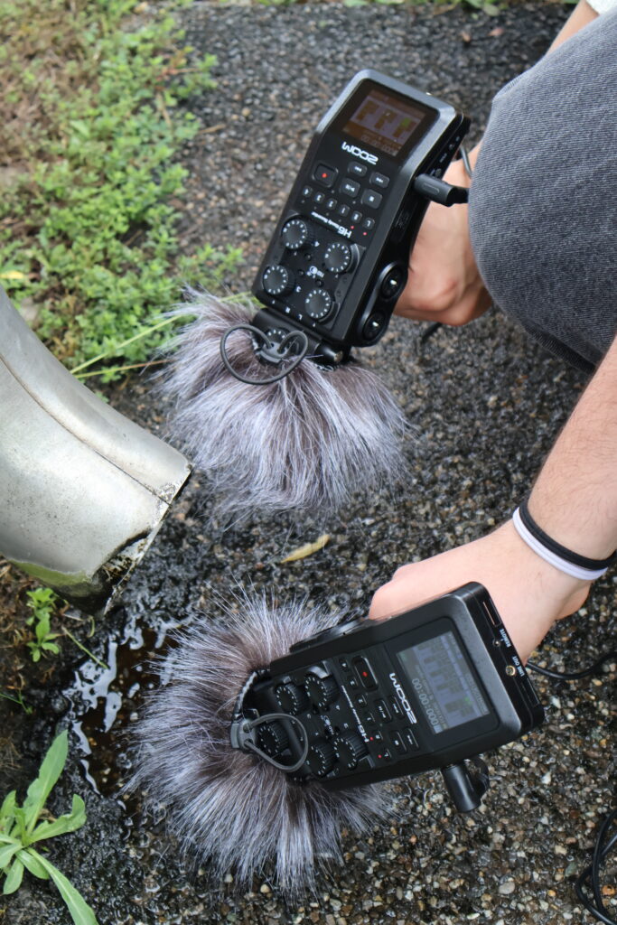 Zwei Personen halten Aufnahmegerät und vor ein Metalrohr.