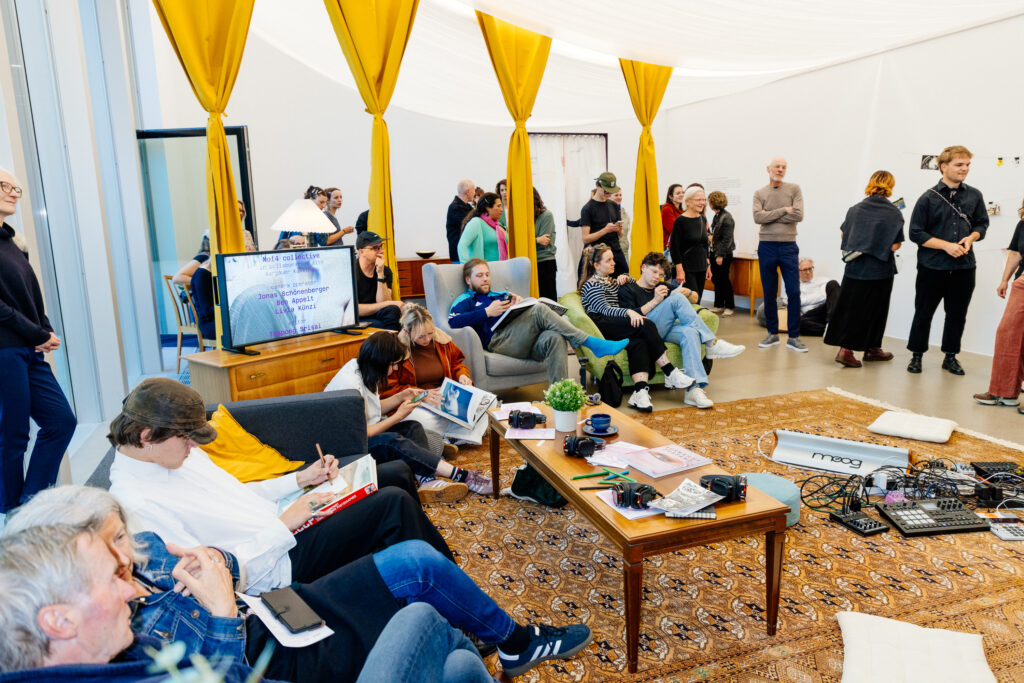 Personen sitzen auf Sofas und auf dem Boden um einen Salontisch auf einem grossen, gemusterten Teppich. Die Personen lesen in Büchern oder schreiben. Im Hintergrund sind gelbe, zugebundene Vorhänge sowie viele stehende und miteinander sprechende Personen zu sehen.