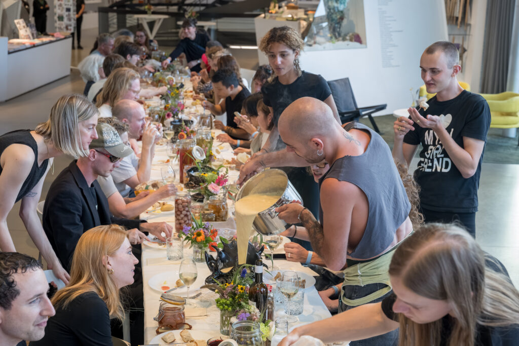 Essende Personen sitzen an einem langen, in den Raum laufenden, mit Blumen, Getränken und Essen vollgestellten Tisch. Eine Person giesst Fondukäse in einen Keramikbehälter.