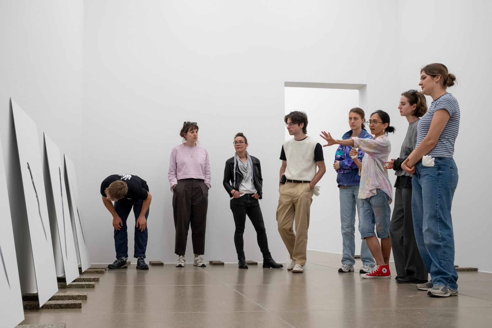 Personen stehen in einem Museum vor an die Wand gelehnten und am Boden stehenden Bildern und diskutieren gemeinsam. Eine Person neigt sich einem der Bilder näher zu.