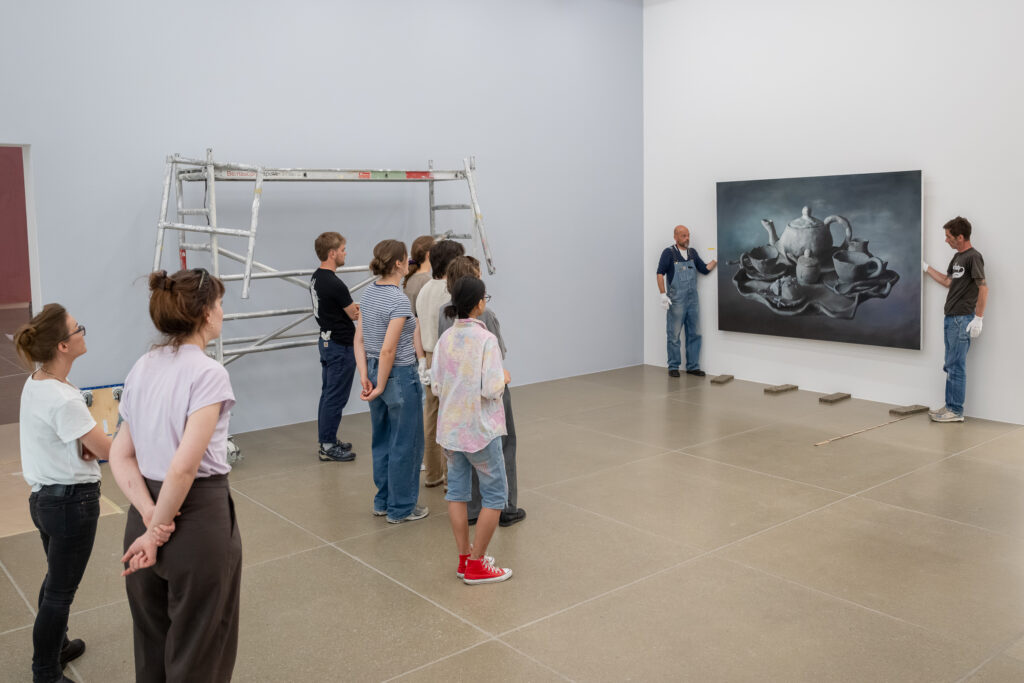Personen schauen in einem Museum zu zwei Personen, die mit weissen Handschuhen ein Gemälde vor einer Wand halten.