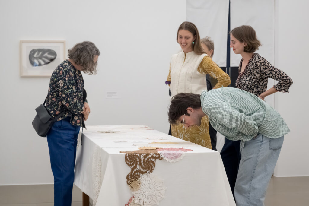Personen stehen um einen Tisch und betrachten das Tischtuch sowie die gehäkelten Untersetzer darauf.