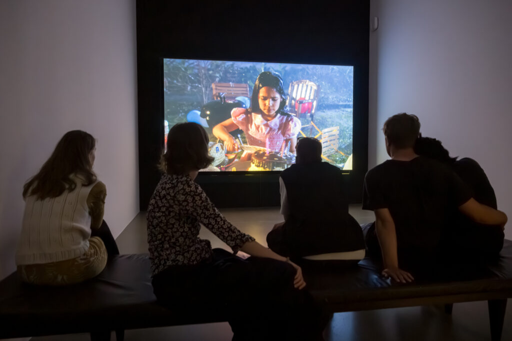 Personen sitzen in einem dunklen Raum auf einer Bank vor einem an die Wand projizierten Video.