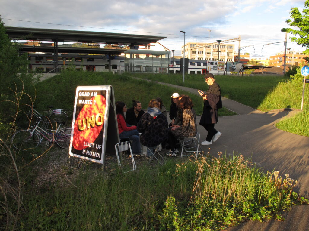 Eine Gruppe Menschen sitzt vor einem Bahnhof auf Klappstühlen im Kreis, und daneben steht ein Klappschild mit einem Plakat, das zum Überbrücken von Wartezeiten mit UNO spielen einlädt.