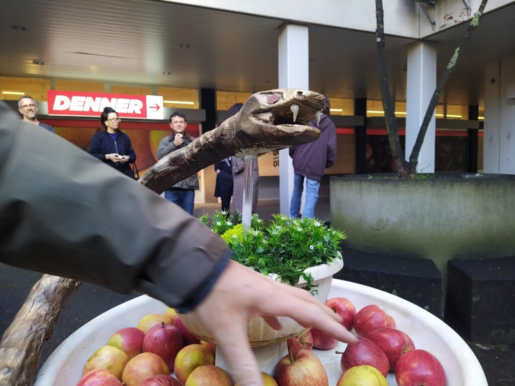 Eine Hand greift in eine Schale mit Äpfeln um eine Topfpflanze. Eine Schlage aus braun und golden angespraytem Material geschnitzte Schlange schwebt oberhalb der Apfelschale.