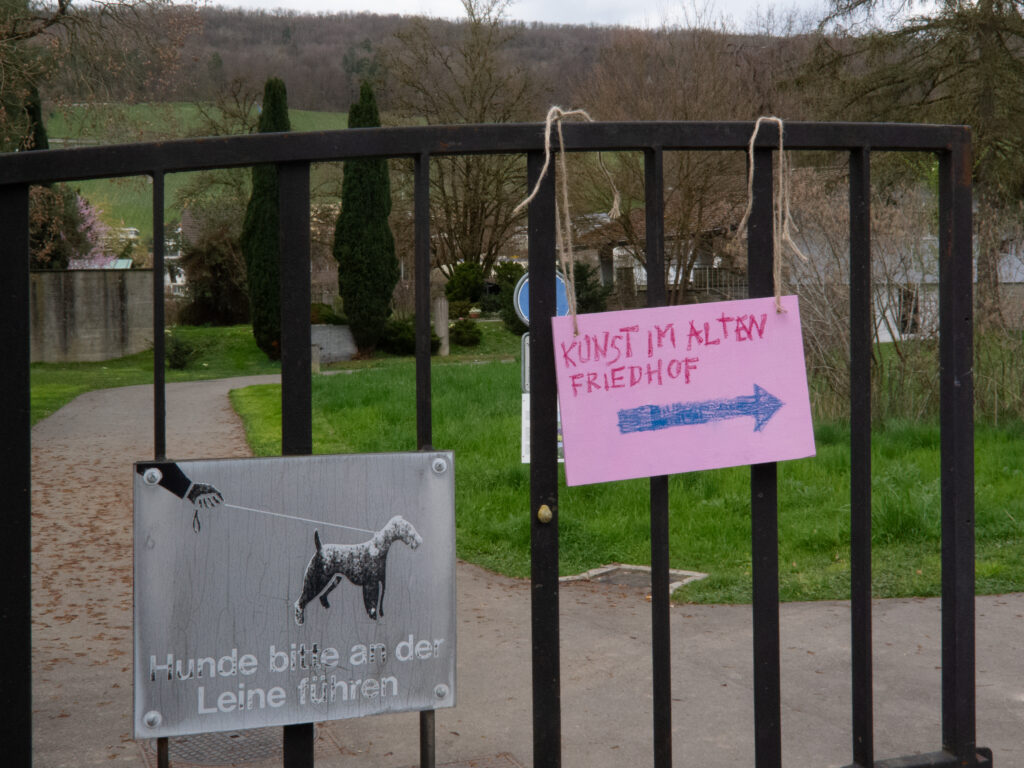 Ein Haag vor einem Friedhof mit zwei Schildern. Eines sagt, dass Hunde an der Leine geführt werden sollen, und auf dem anderen wird handschriftlich mit einem Pfeil auf eine Kunstausstellung auf dem Friedhof aufmerksam gemacht.