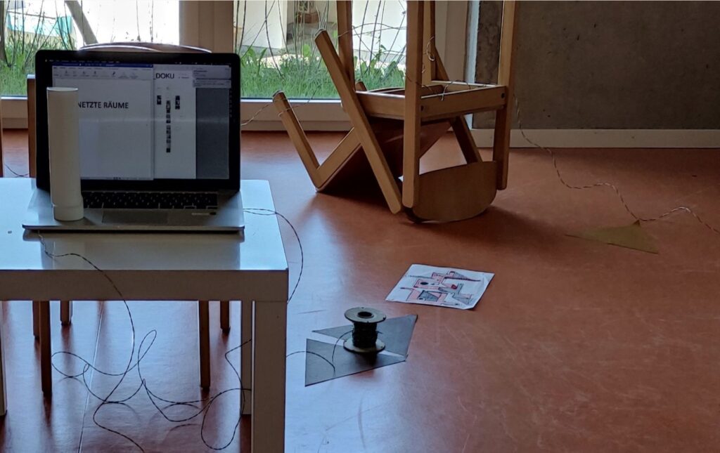 Zwei Stühle liegen ineinander verhakt am Boden. Auf einem Tisch steht ein Laptop. Die Gegenstände im Raum sind mit einem Kabel verbunden.