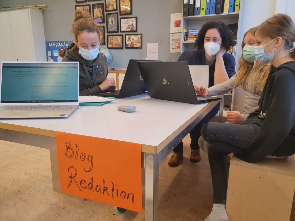 Schülerinnen sitzen mit Laptops an einem Tisch. Am Tisch klebt ein orangener Zettel mit der Aufschrift Blog Redaktion.