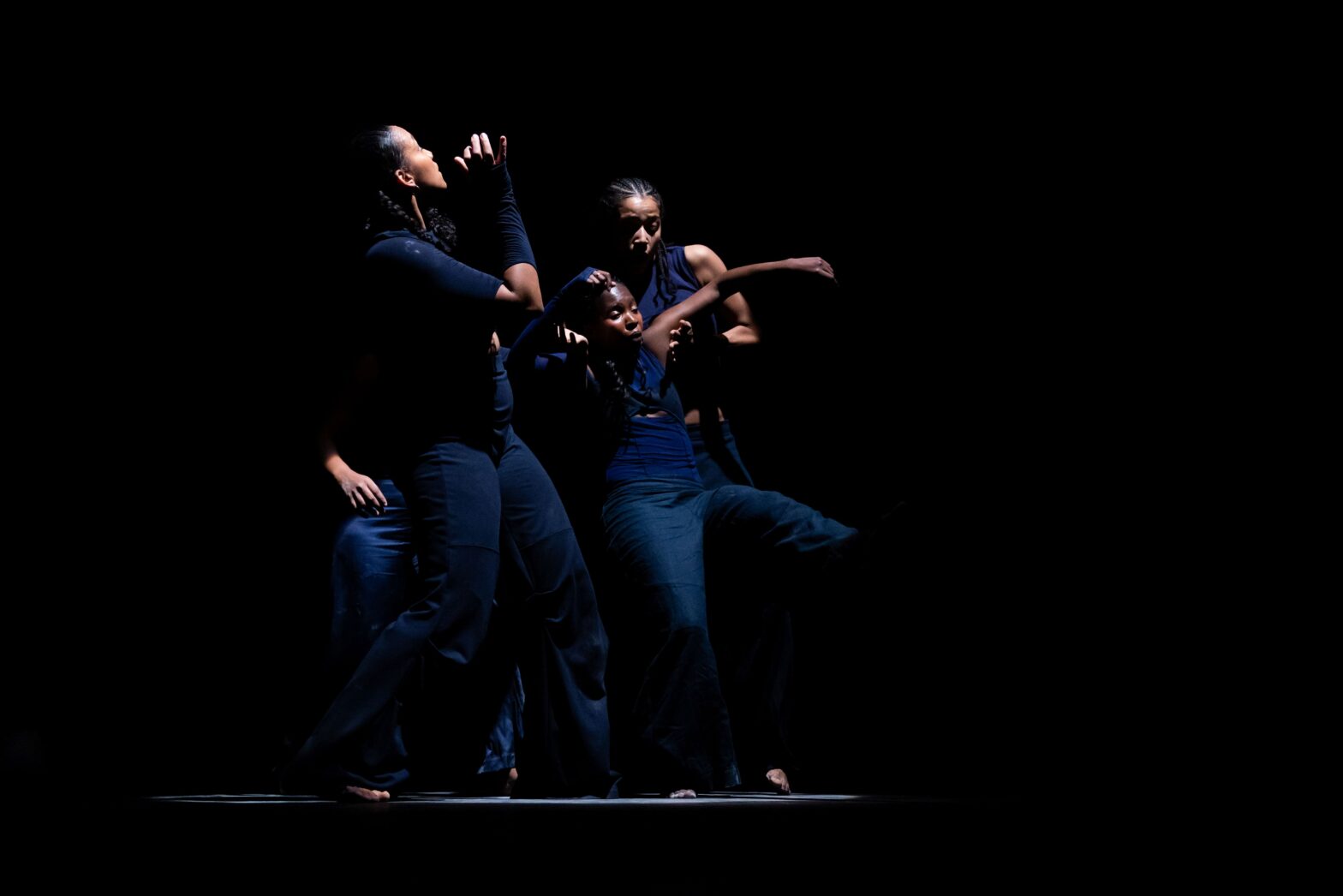 Vier Frauen in blauer Kleidung tanzen in einem Lichtstrahl auf der Bühne, während es rundherum dunkel ist.