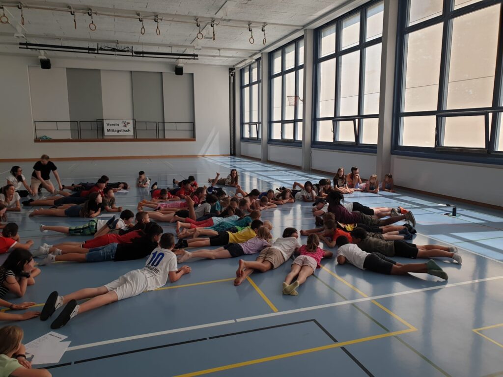 viele Schülerinnen und Schüler liegen in einer Turnhalle auf dem Boden