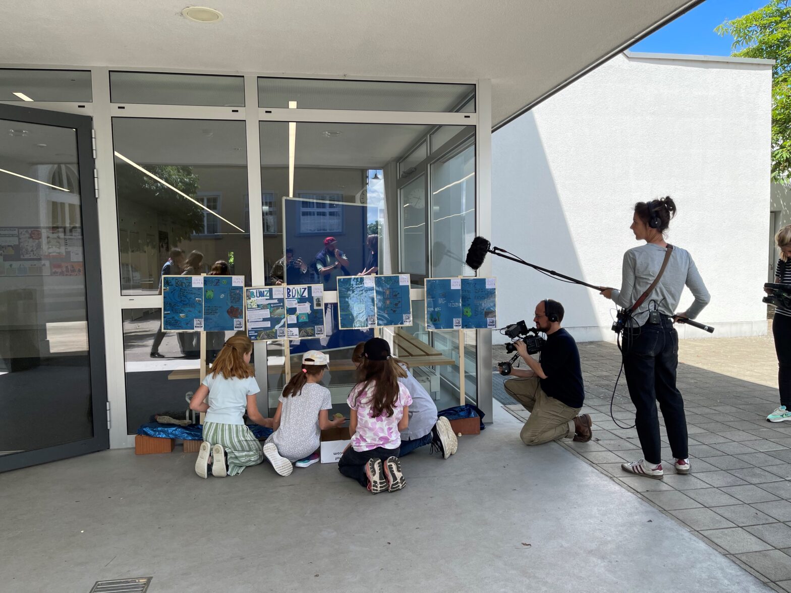 Schülerinnen und Schüler werden arbeiten draussen an Plakaten und werden von einem Kamerateam gefilmt.