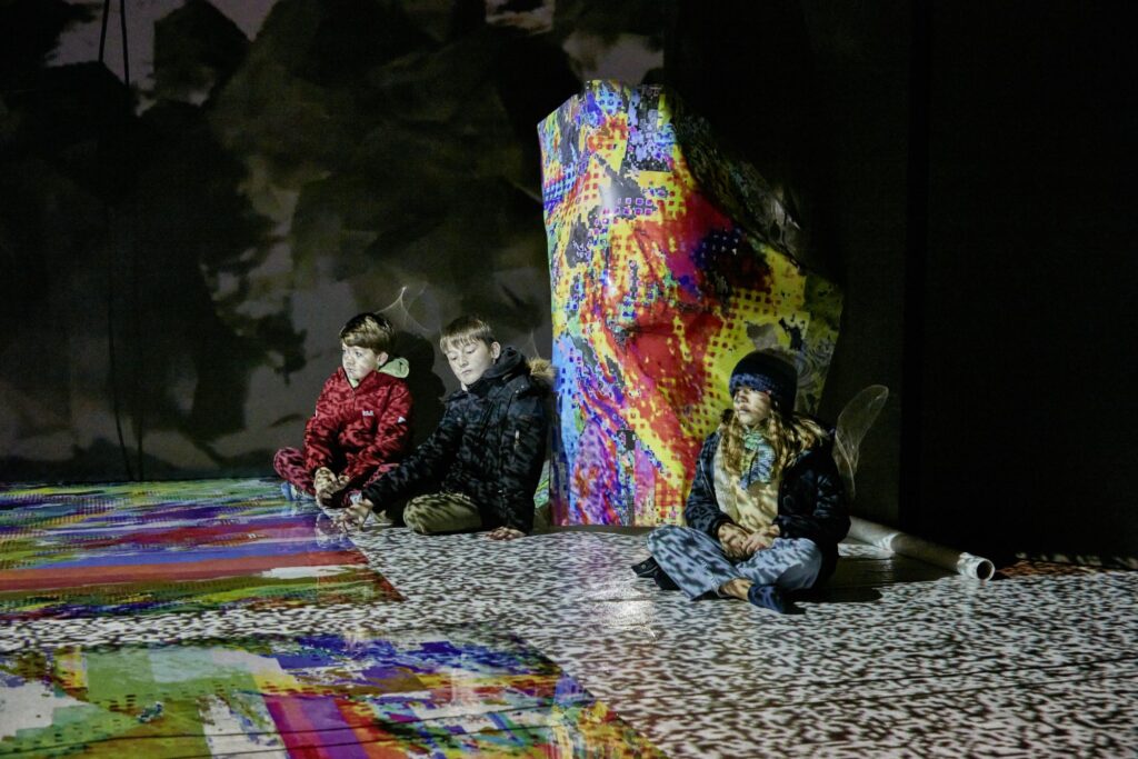 Drei Kinder sitzen in einem dunklen Raum mit bunten Elementen.